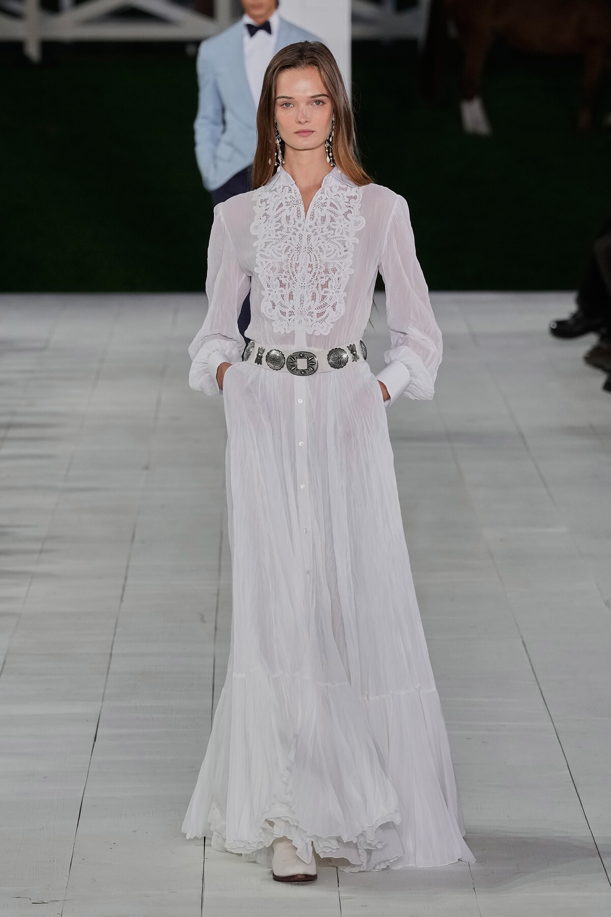 Model on a runway wearing a sheer white maxi dress with lace bib, belted at the waist with a large silver belt, hands in pockets.