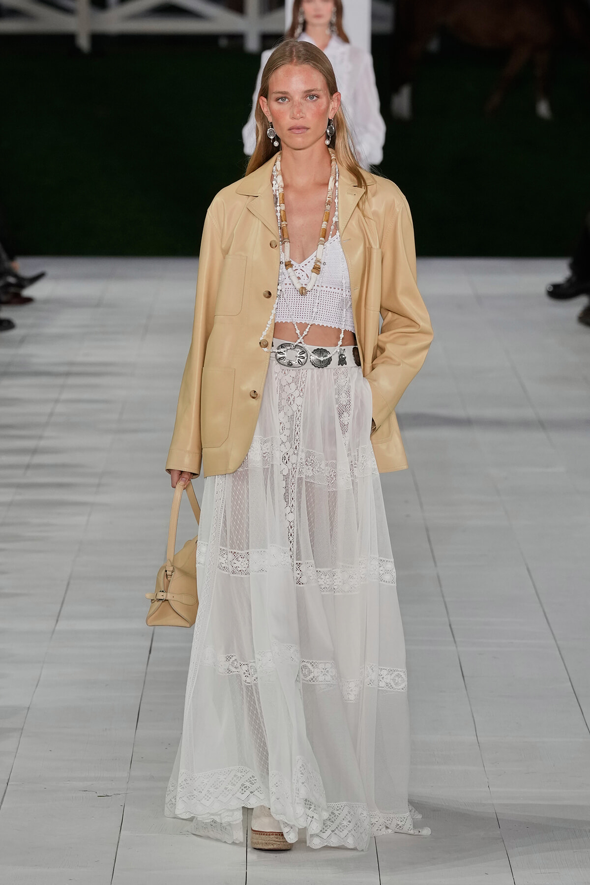 Model on a runway wearing a tan blazer over a white crochet crop top and tiered lace maxi skirt, accessorized with a beaded necklace, silver belt, earrings, and a small tan handbag.