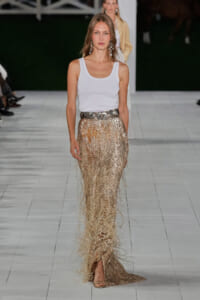 Model walking the runway in a white tank top and gold fringe sequin skirt with a bold belt and statement earrings.