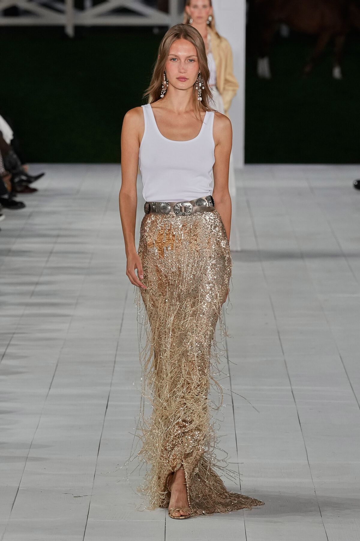 Model walking the runway in a white tank top and gold fringe sequin skirt with a bold belt and statement earrings.