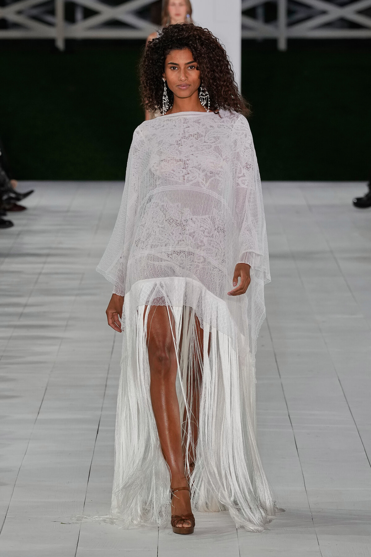 Model walks the runway in a white lace sheer dress with a long fringe train and statement chandelier earrings.
