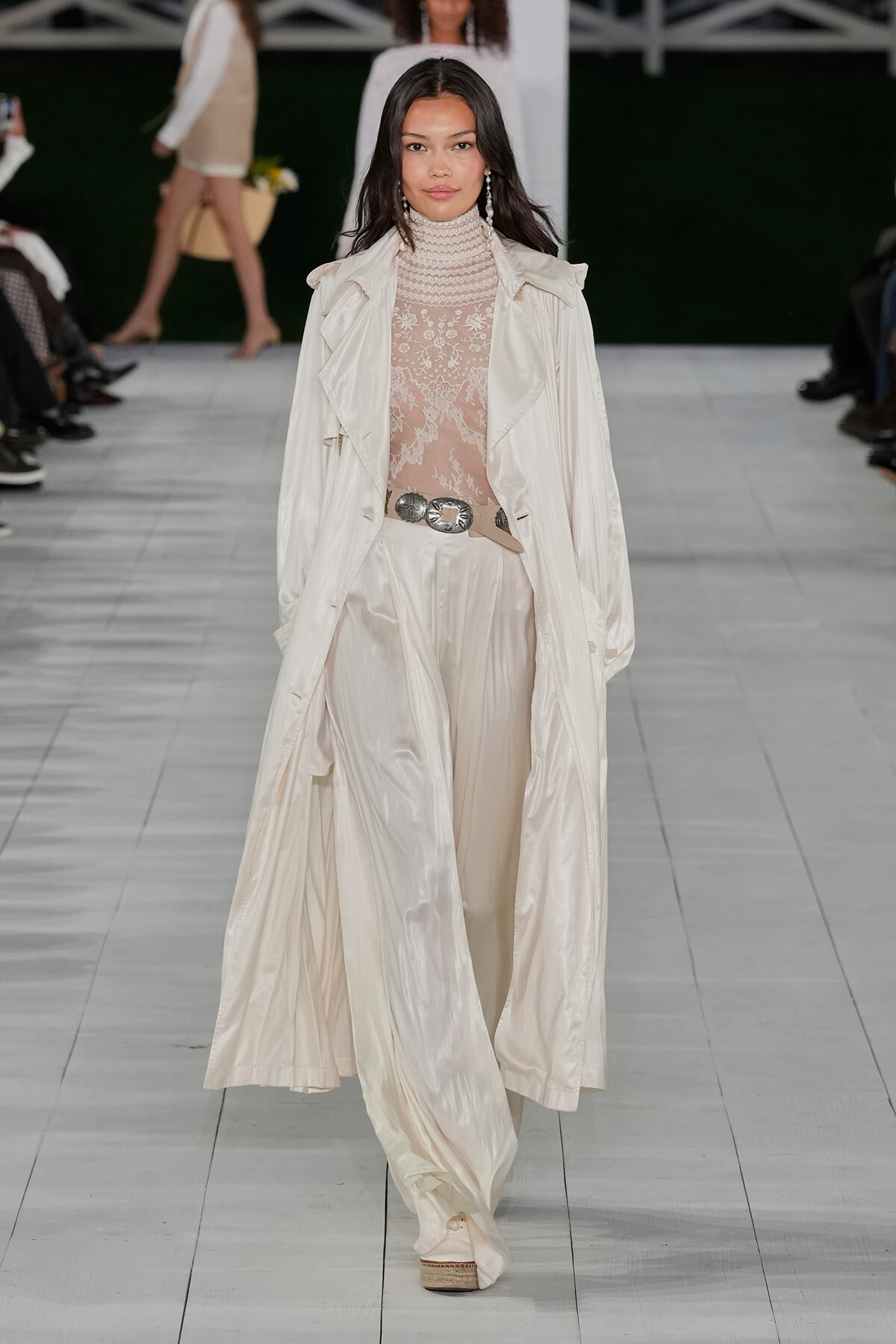 Model walks the runway in an ivory trench coat over a sheer lace turtleneck and wide-leg ivory pants, with a decorative belt.