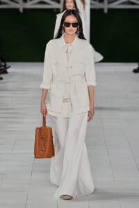 Model on a runway wearing an ivory utility jacket tied at the waist with matching wide-leg pants, accessorized with brown sunglasses and a tan leather tote.