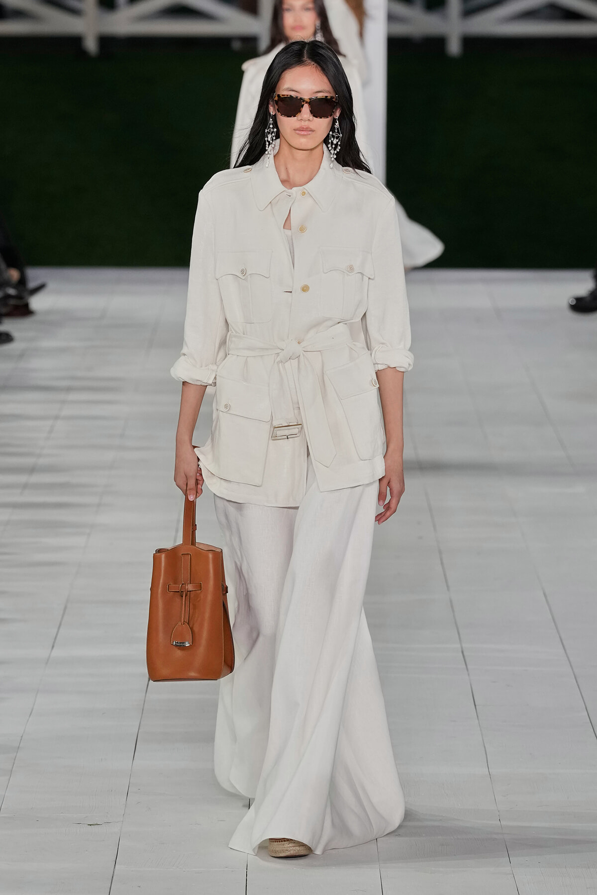 Model on a runway wearing an ivory utility jacket tied at the waist with matching wide-leg pants, accessorized with brown sunglasses and a tan leather tote.