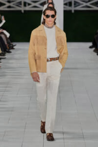 Male model on a runway wearing a tan suede fringe jacket over a cream knit turtleneck with white pants and brown shoes.