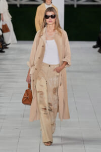Model walking a fashion runway in a beige trench over a white top and glittery ripped pants, wearing dark sunglasses and carrying a tan handbag.