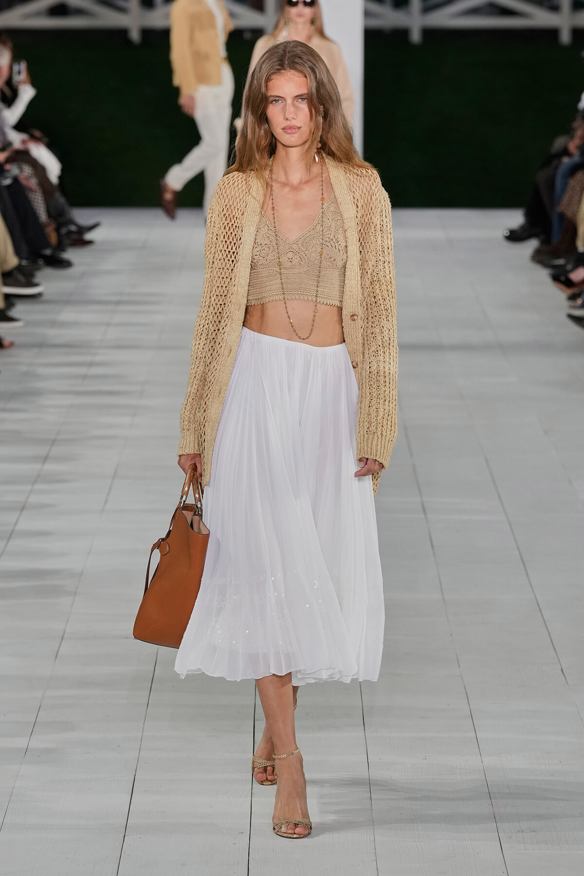 Runway model wearing a beige crochet cardigan over a lace bralette and white flowy midi skirt, carrying a tan leather tote.