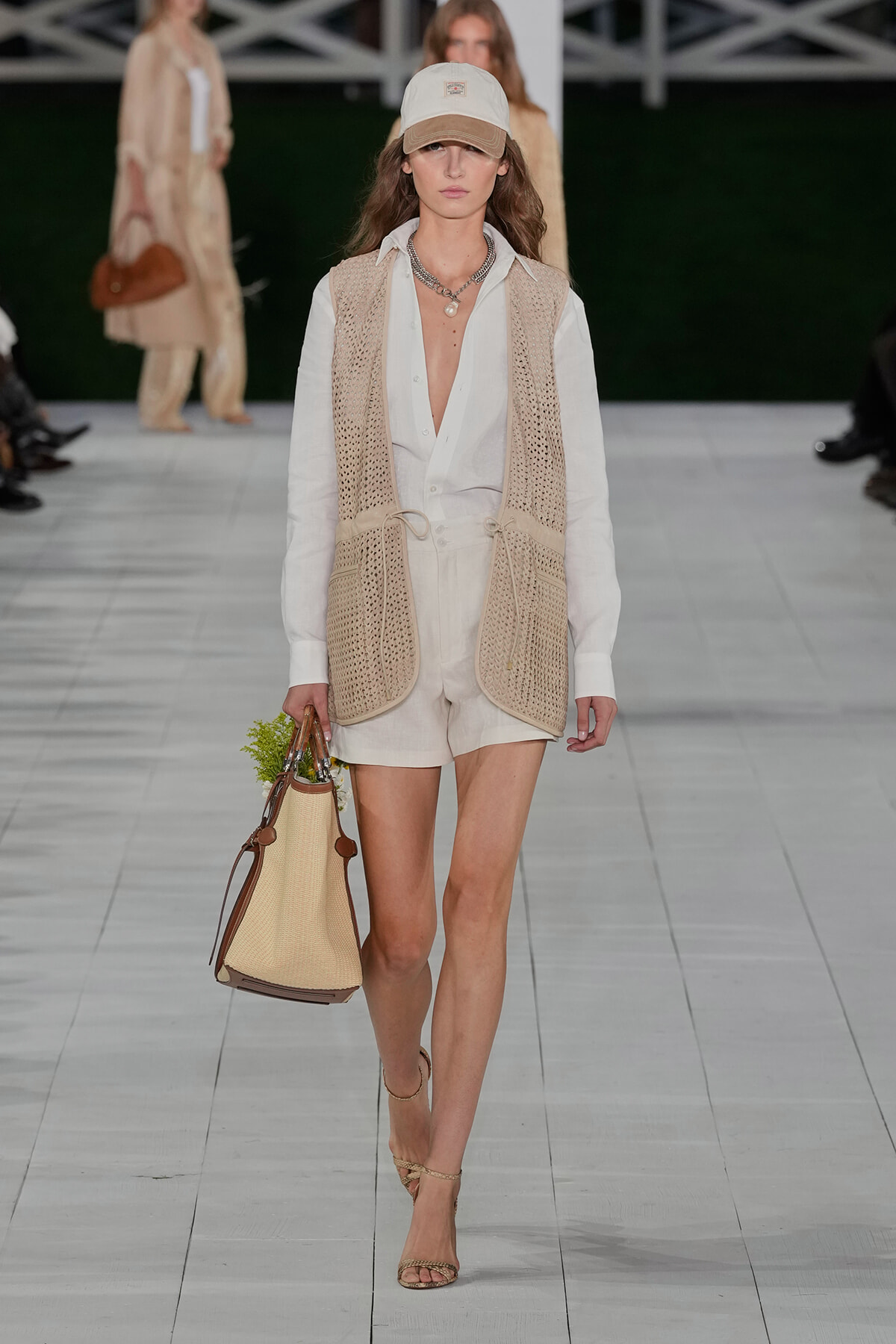 Model on a runway wearing a white shirt, beige knit vest, and light shorts, carrying a straw tote bag and wearing a beige cap.