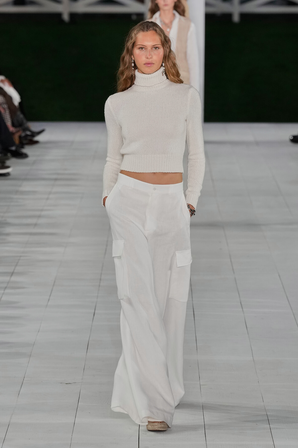 Female model walking the runway in a cream knit turtleneck and loose white cargo pants with hands in pockets.