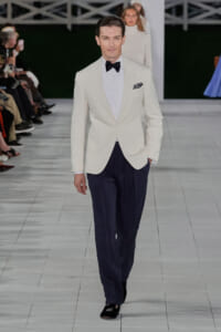 Male model on a runway wearing a white dinner jacket, black bow tie, white shirt, navy trousers, and black loafers with a pocket square in the lapel.