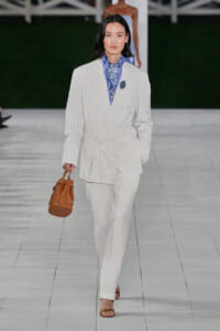 Model walks the runway in a white pinstripe, double-breasted blazer with matching trousers, blue patterned scarf, and a brown bucket bag with tan sandals.