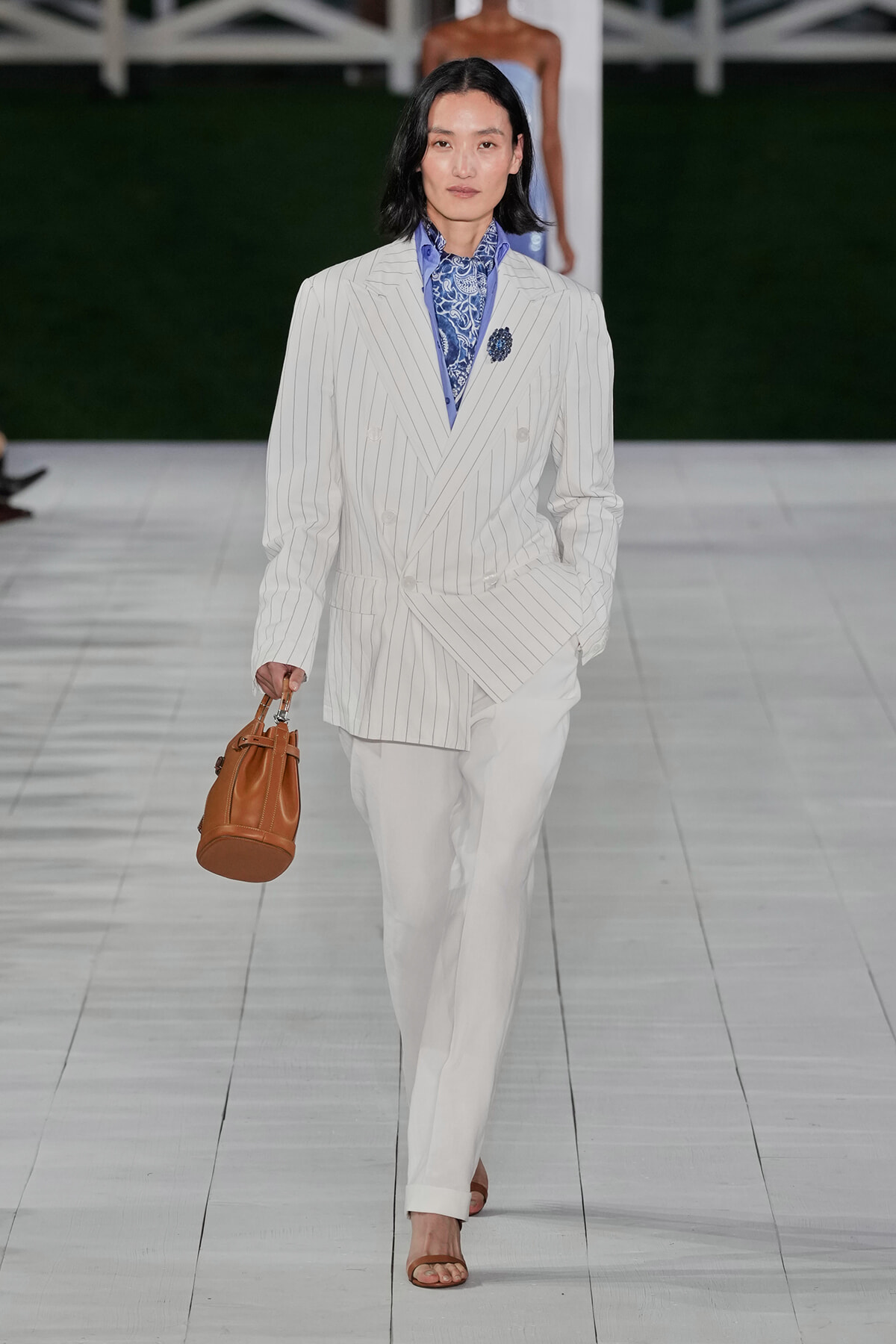 Model walks the runway in a white pinstripe, double-breasted blazer with matching trousers, blue patterned scarf, and a brown bucket bag with tan sandals.