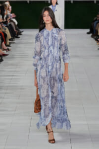 Female model walks a runway wearing a blue lace maxi dress with long sleeves, carrying a tan leather bag.