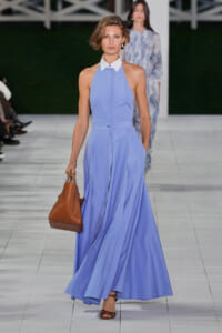 Model walks the runway in a light blue halter dress with a white collar, carrying a brown handbag and wearing brown sandals.