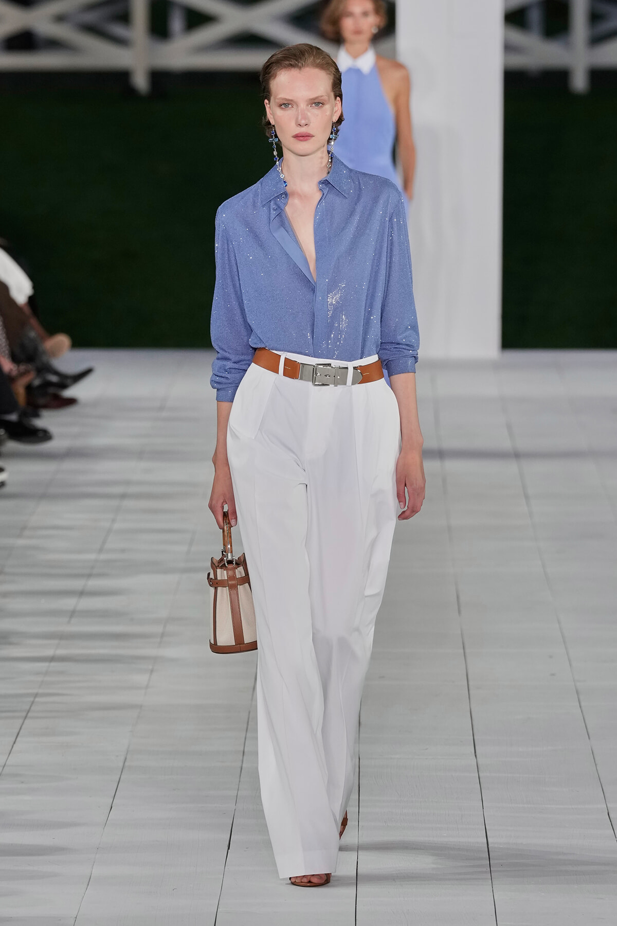 Fashion model on a runway wearing a light blue, sequined shirt tucked into white wide-leg trousers with a brown belt and matching brown-trim bucket bag.