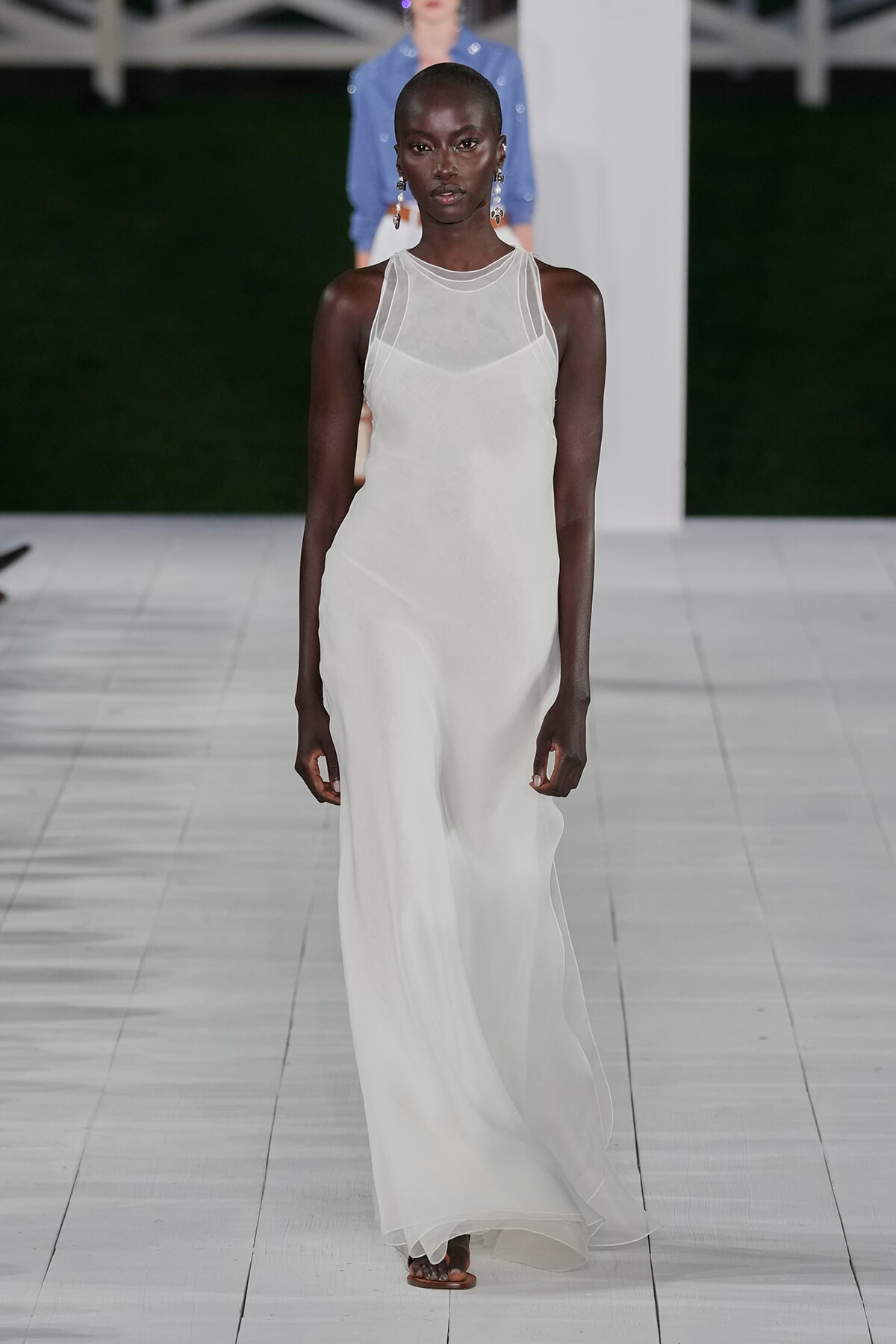 Model walks a white, floor-length gown with a sheer, sleeveless yoke and subtle layering on a bright runway.
