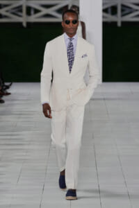 Male model walks a runway in a white suit, lavender striped shirt, patterned tie, and dark sunglasses.