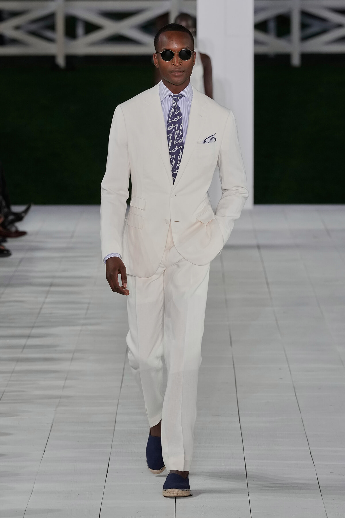 Male model walks a runway in a white suit, lavender striped shirt, patterned tie, and dark sunglasses.