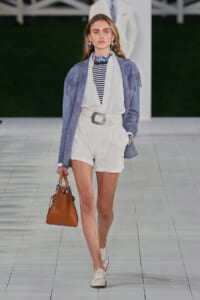 Runway model wearing a light blue fringe suede jacket over a white belted romper, striped top, blue scarf, tan handbag, and cream Oxford shoes.