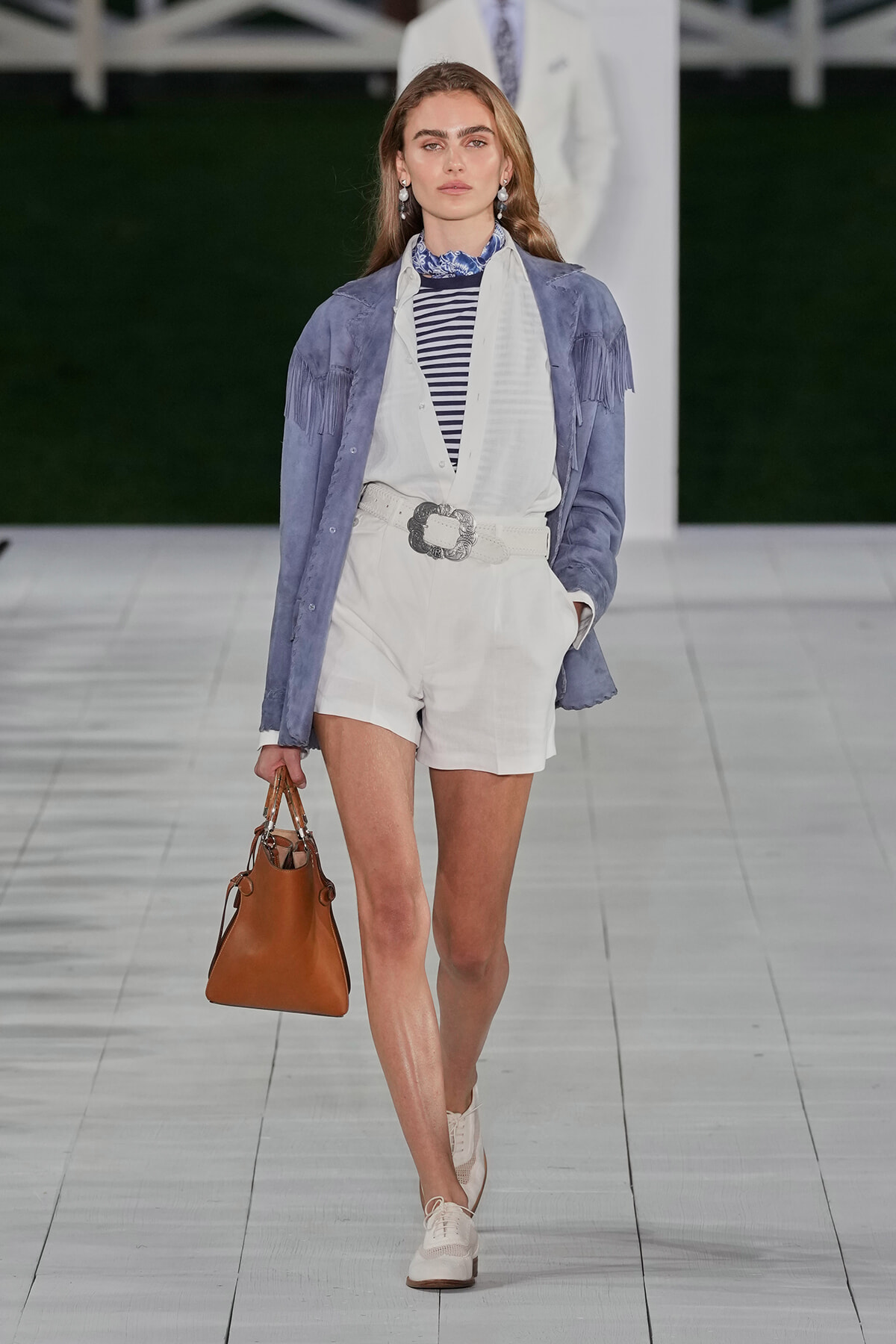 Runway model wearing a light blue fringe suede jacket over a white belted romper, striped top, blue scarf, tan handbag, and cream Oxford shoes.