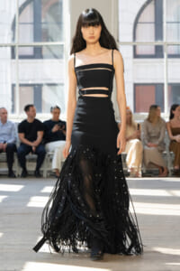 Model in a black cutout evening gown with fringe panels walks a runway indoors, audience in background.