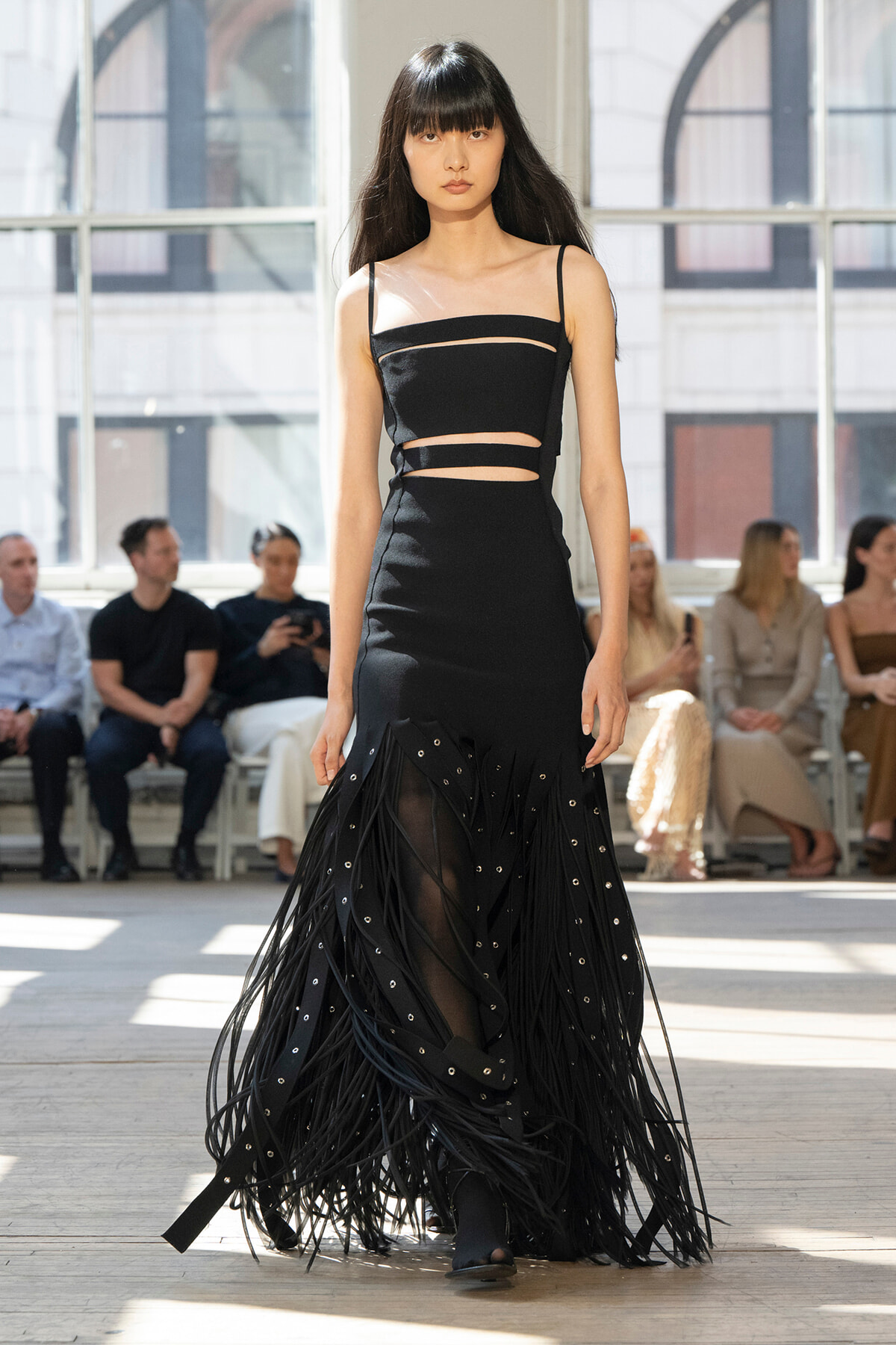 Model in a black cutout evening gown with fringe panels walks a runway indoors, audience in background.
