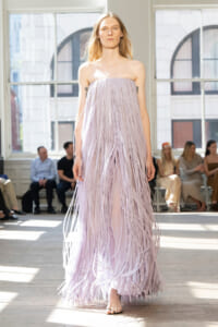Model walking a lavender fringe strapless gown down a runway, audience watching in a bright studio.
