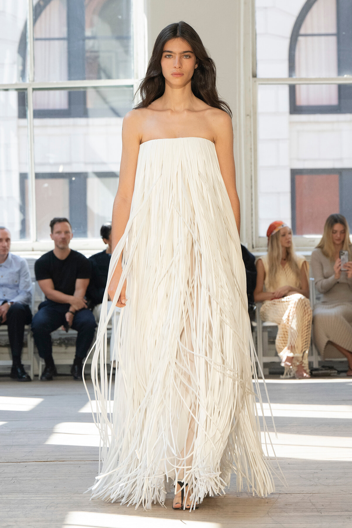 Model walking the runway in a strapless white fringe gown; audience seated in the background.