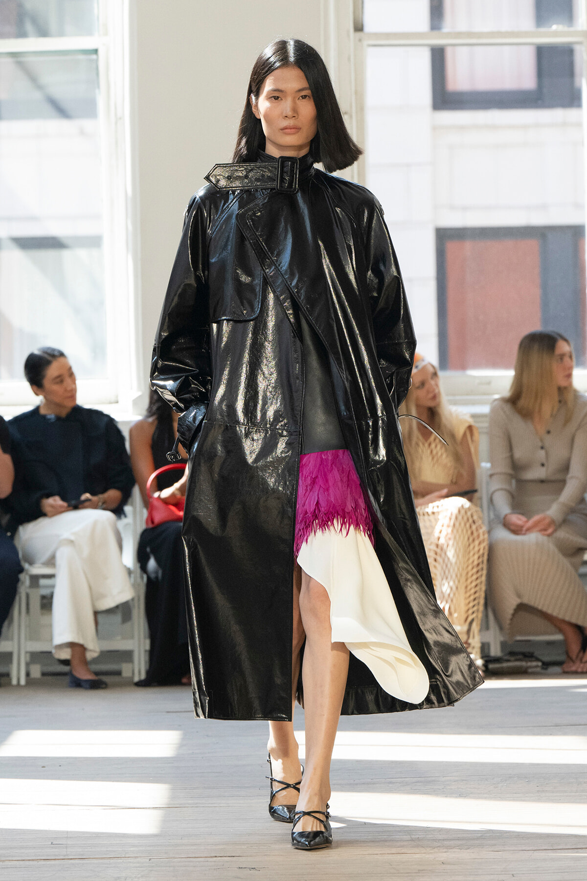 Model walking the runway in a shiny black trench coat with a high collar; magenta feathered hem peeks from beneath, audience seated in the background.