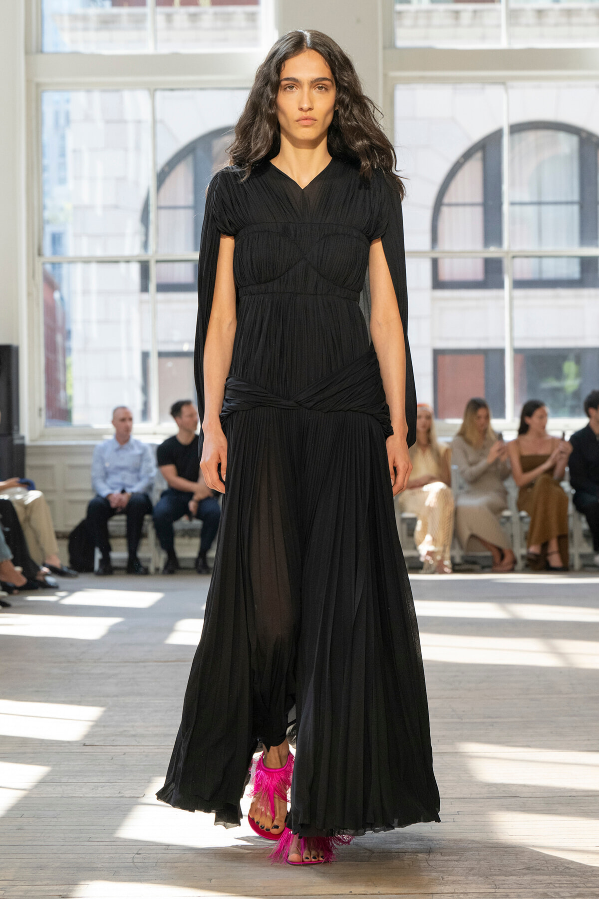 Model walks the runway in a long black pleated gown with cape sleeves and bright pink feathered sandals; audience seated in the background.