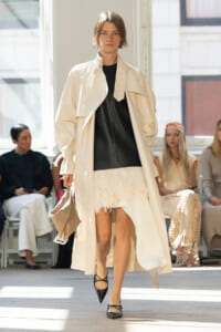 Model walking the runway in a cream trench coat over a black top and a feathered white mini skirt, with audience in the background.