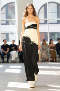 Model walking a runway in a strapless cream-and-black color-block dress with a black waist band, white boots, and an audience in the background.