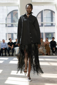 Model walking a fashion runway in a black oversized button-down shirt paired with a long black fringe skirt and silver pointed heels; audience seated in the background.