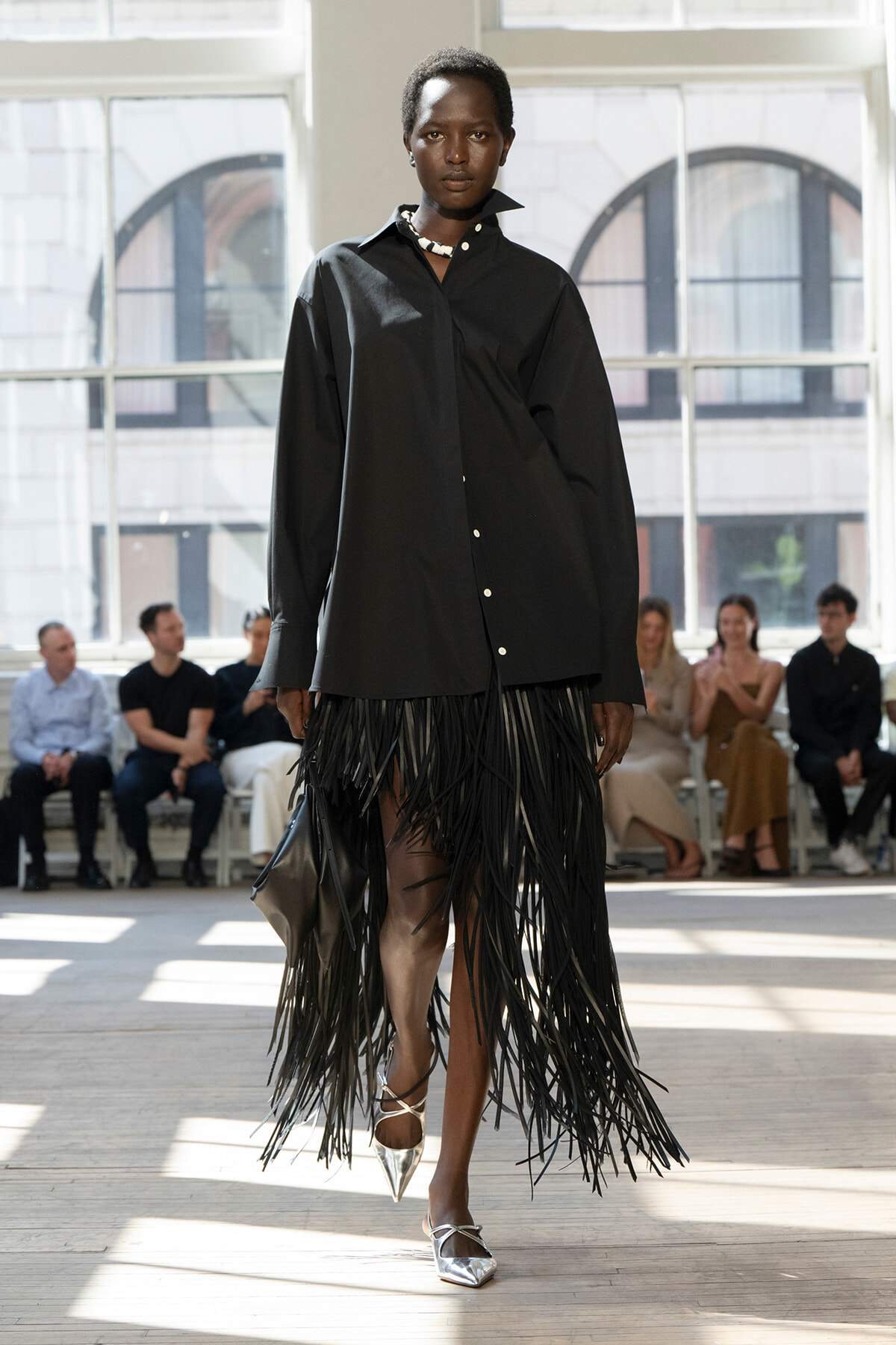 Model walking a fashion runway in a black oversized button-down shirt paired with a long black fringe skirt and silver pointed heels; audience seated in the background.
