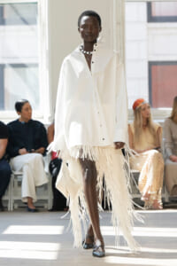 Model walks the runway in an oversized white shirt with fringe trim at the hem, accessorized with a beaded necklace.
