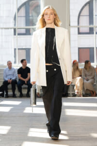 Model on a fashion runway wearing a white blazer over a black top and wide-leg black pants, holding a metallic clutch as she walks.