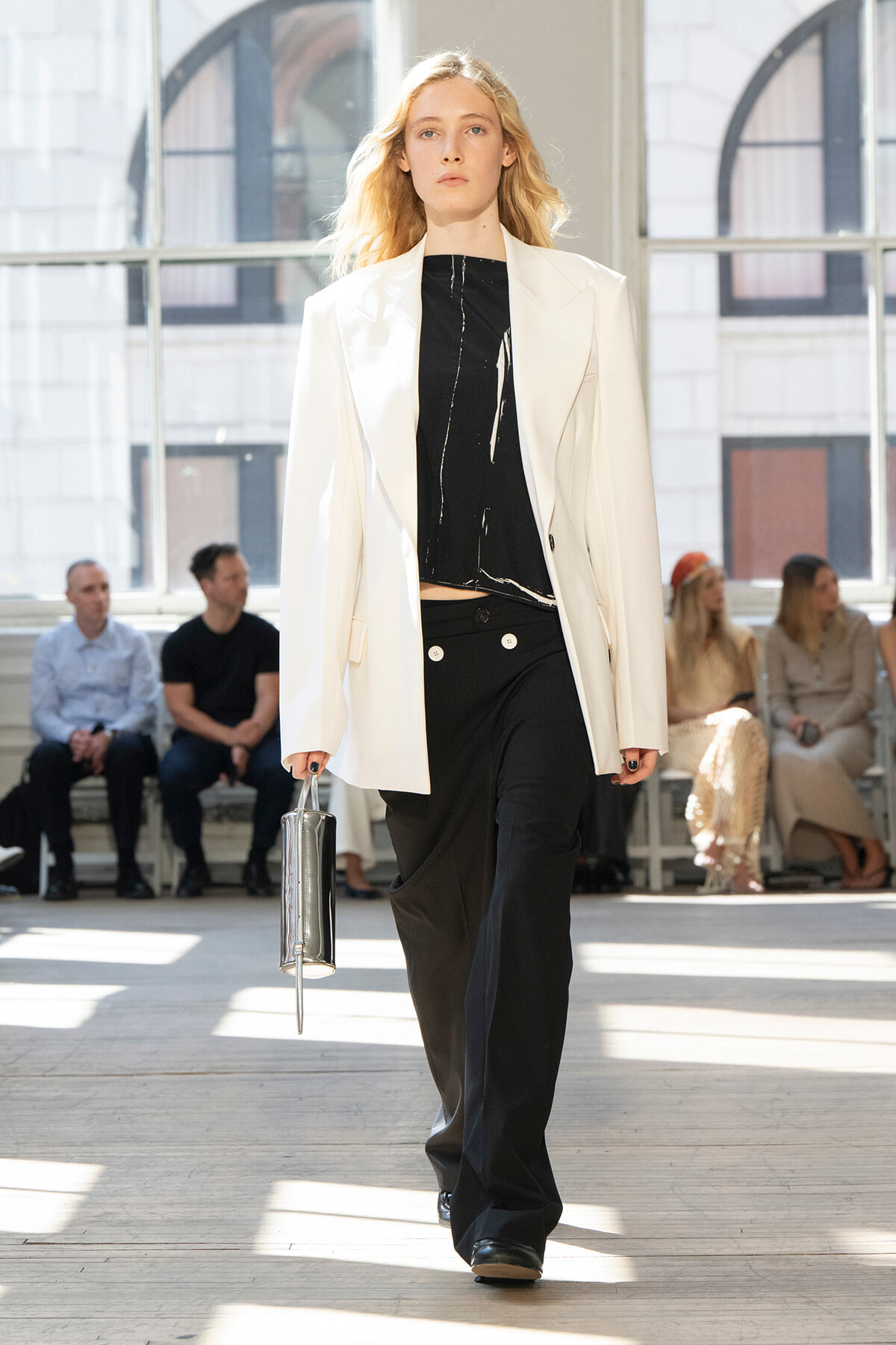 Model on a fashion runway wearing a white blazer over a black top and wide-leg black pants, holding a metallic clutch as she walks.