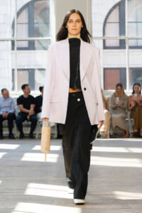 Model on a runway wearing a pale pink blazer over a black zip-front top and wide-leg black pants, carrying a small beige handbag.