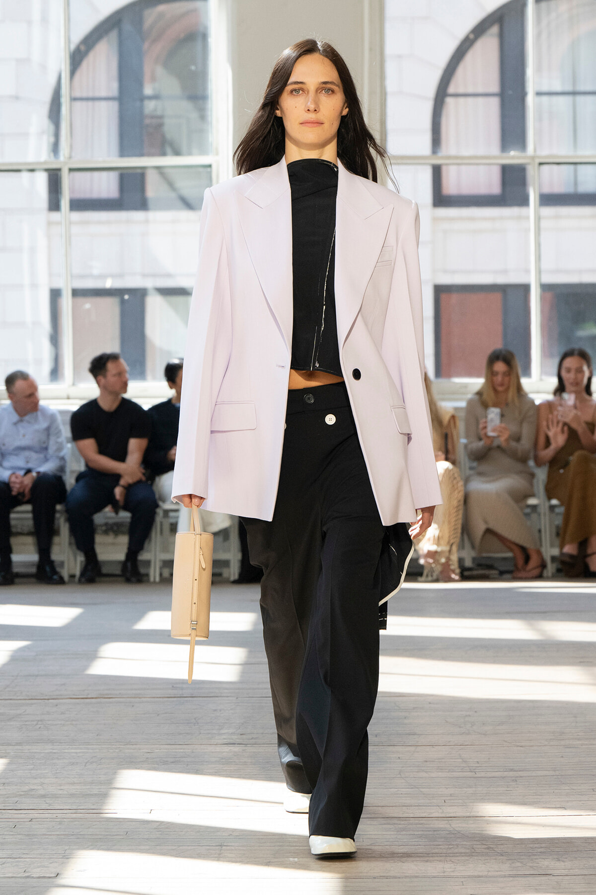 Model on a runway wearing a pale pink blazer over a black zip-front top and wide-leg black pants, carrying a small beige handbag.