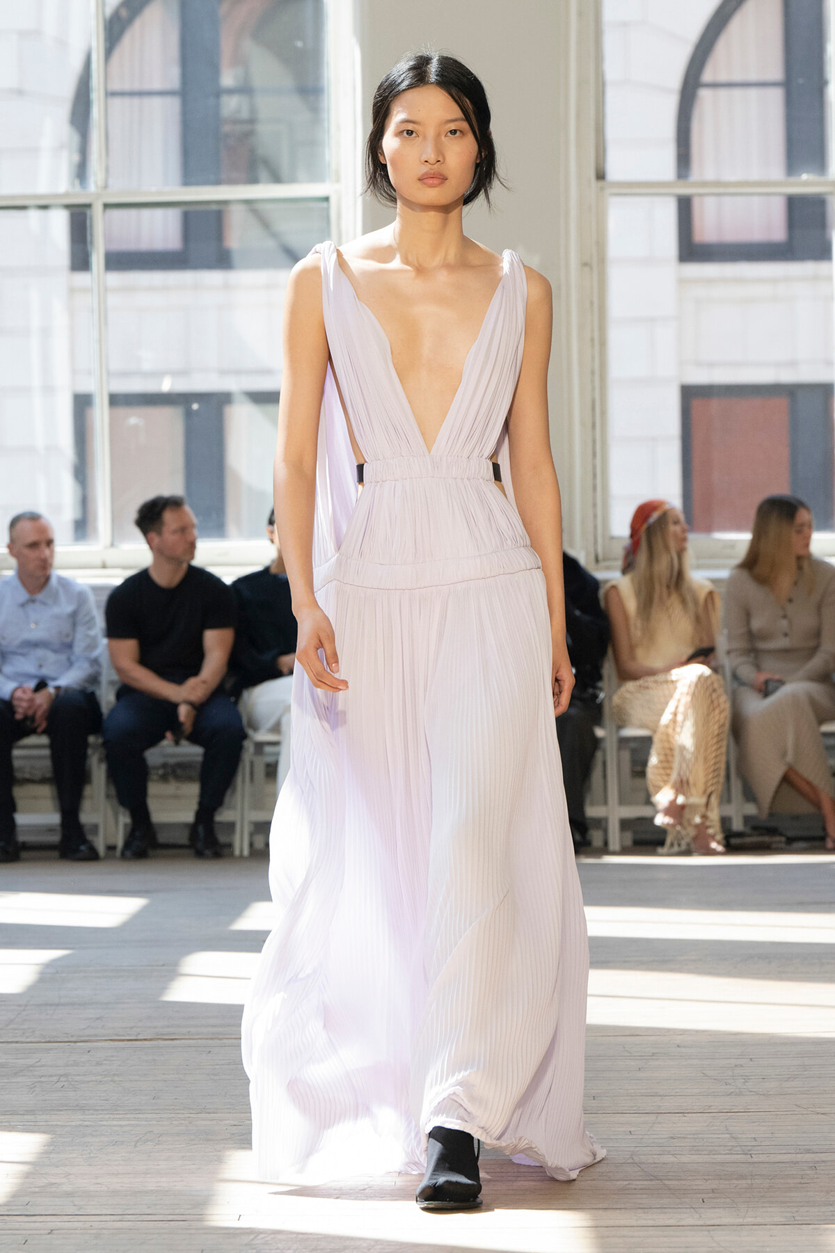 Model walks the runway in a light lavender pleated gown with a deep plunging V neckline; audience sits in the background.