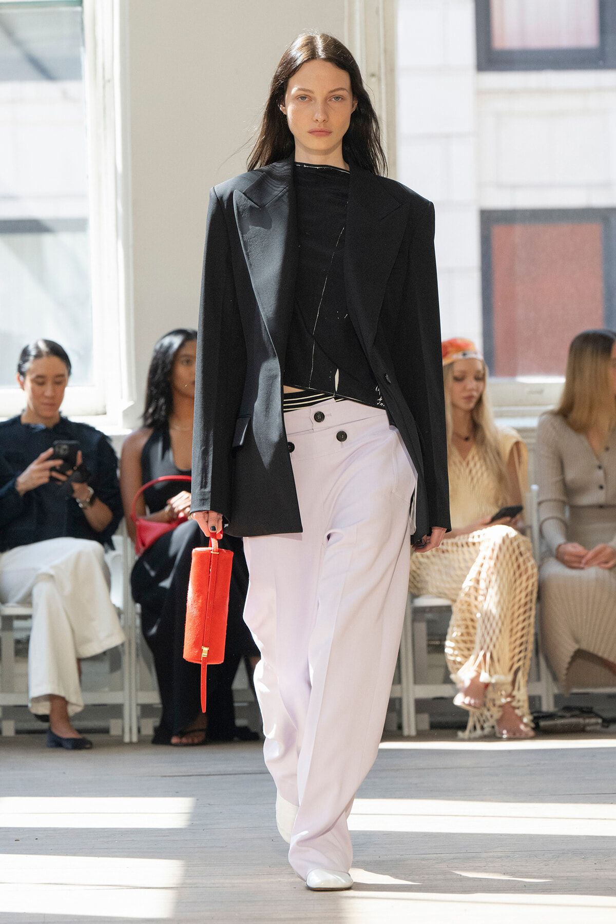 Model walking runway in oversized black blazer, black top, white wide-leg pants, carrying a small red handbag.