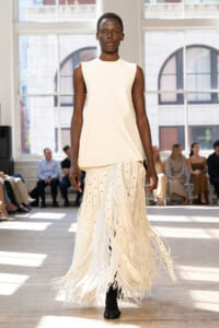 Model walks a runway in an ivory sleeveless top paired with a long fringe skirt; bright studio setting with seated audience in background.