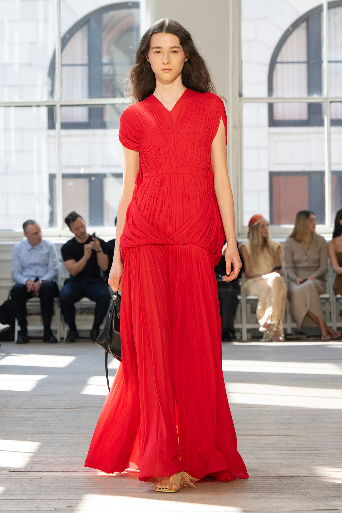 Model walks a runway wearing a bright red, pleated gown with a wrapped bodice and flowing skirt, holding a black bag; audience seated in a sunlit studio behind her.