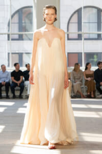 Model walks the runway in a strapless cream pleated gown with a plunging V neckline; audience seated in a bright studio behind her.