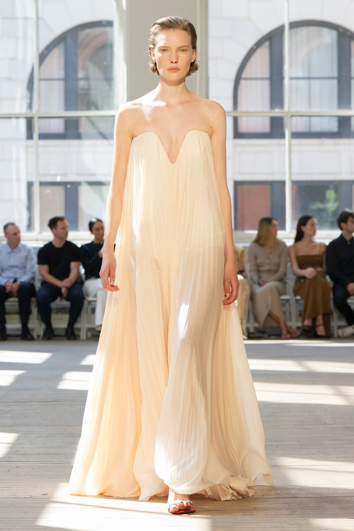 Model walks the runway in a strapless cream pleated gown with a plunging V neckline; audience seated in a bright studio behind her.