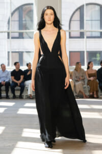 Model walks a runway in a long black pleated gown with a deep V neckline, audience seated along the sides in a sunlit venue.