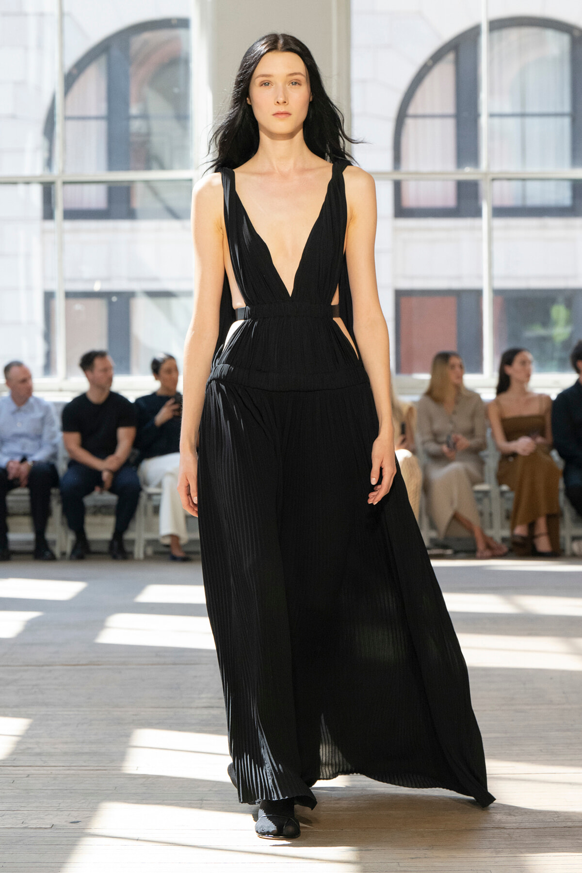 Model walks a runway in a long black pleated gown with a deep V neckline, audience seated along the sides in a sunlit venue.