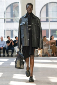 Black model struts down runway in an oversized black leather jacket over a black dress, carrying a large perforated black tote bag.