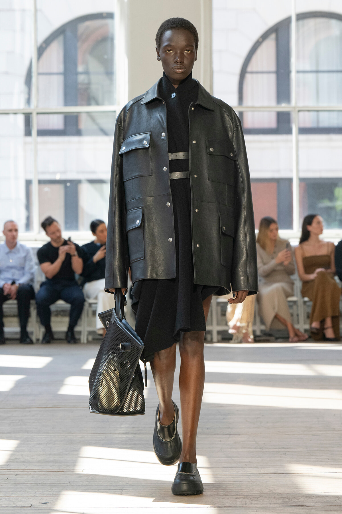 Black model struts down runway in an oversized black leather jacket over a black dress, carrying a large perforated black tote bag.