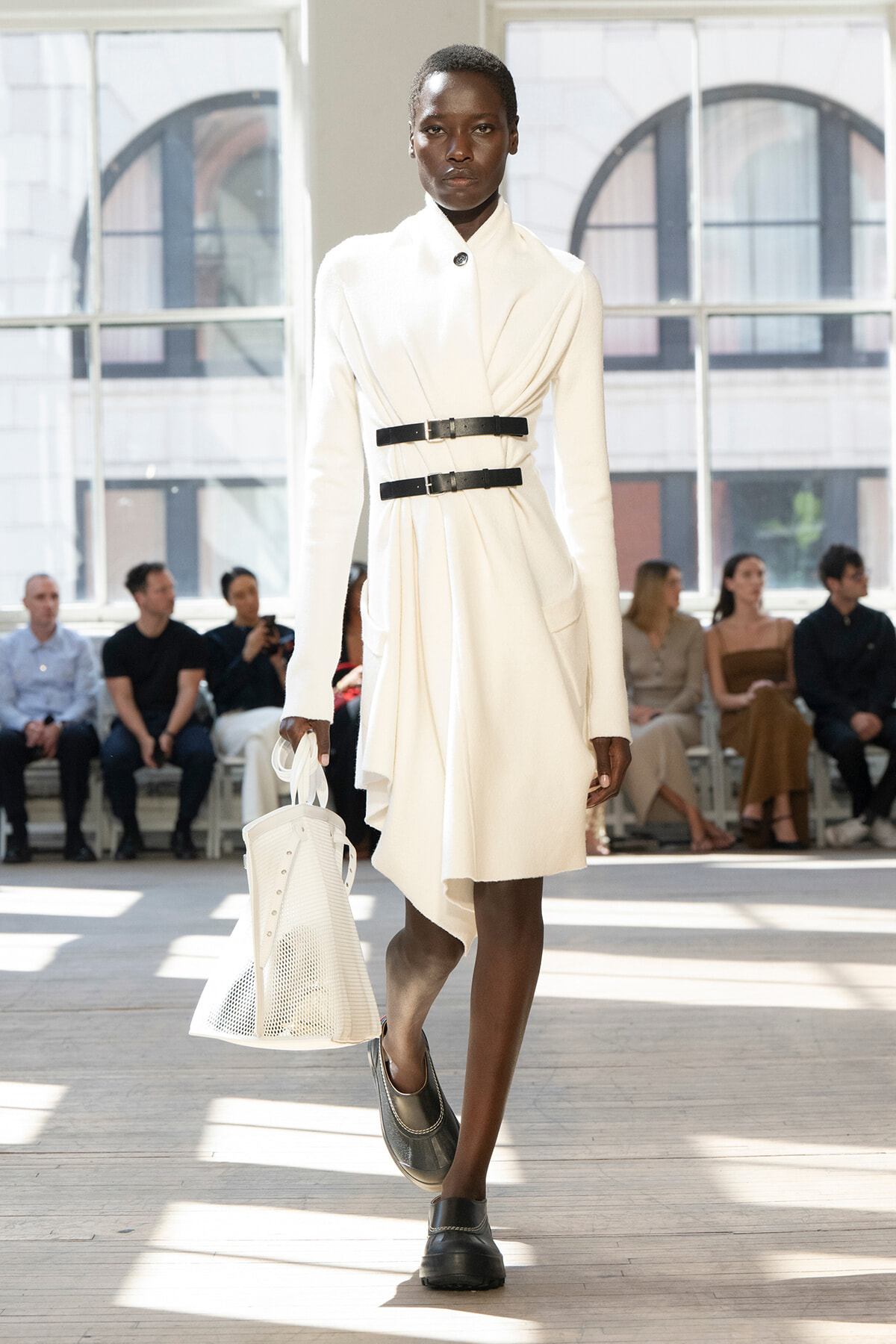 Model on a runway wearing a white draped coat with two black belts, carrying a white perforated tote; audience seated in the background.